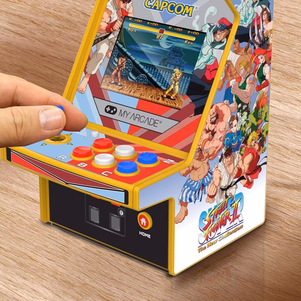 Hand pressing the joystick on a yellow mini arcade cabinet with Street Fighter II artwork and colorful buttons.