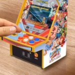 Hand pressing the joystick on a yellow mini arcade cabinet with Street Fighter II artwork and colorful buttons.